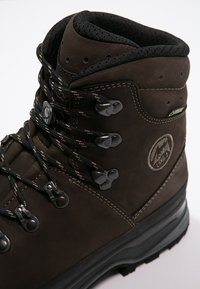 Bota de caminhada em couro castanho com superfície texturizada, biqueira reforçada e ilhós de metal. Apresenta atacadores pretos e um colar almofadado para suporte.