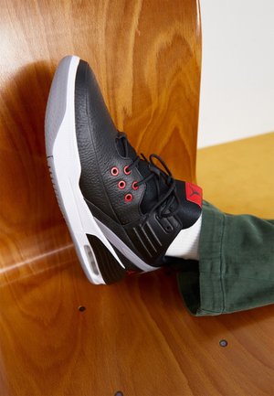 Scarpa da basket bianca e nera con occhielli rossi e logo, indossata con pantaloni verdi, appoggiata allo schienale di una sedia di legno.