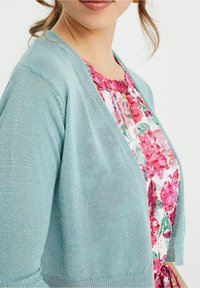 Světle modrý třpytivý cardigan s měkkou texturou, otevřeným předním dílem a žebrovaným lemem, nošený přes květované šaty s růžovými a zelenými odstíny.