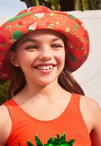 Orange badesuit med grønne palietter, parret med en rød bøttehat med jordbær- og blomstermønstre. Lyse, glade farver.