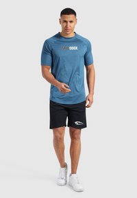 Camiseta atlética azul de manga corta con logo; combinada con pantalones cortos negros que presentan un diseño en blanco; modelo llevando zapatillas blancas.