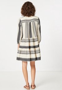 Kvinna med lockigt hår som bär en knälång krämfärgad klänning med svarta geometriska mönster och beigea platta sandaler, stående med ryggen mot oss.