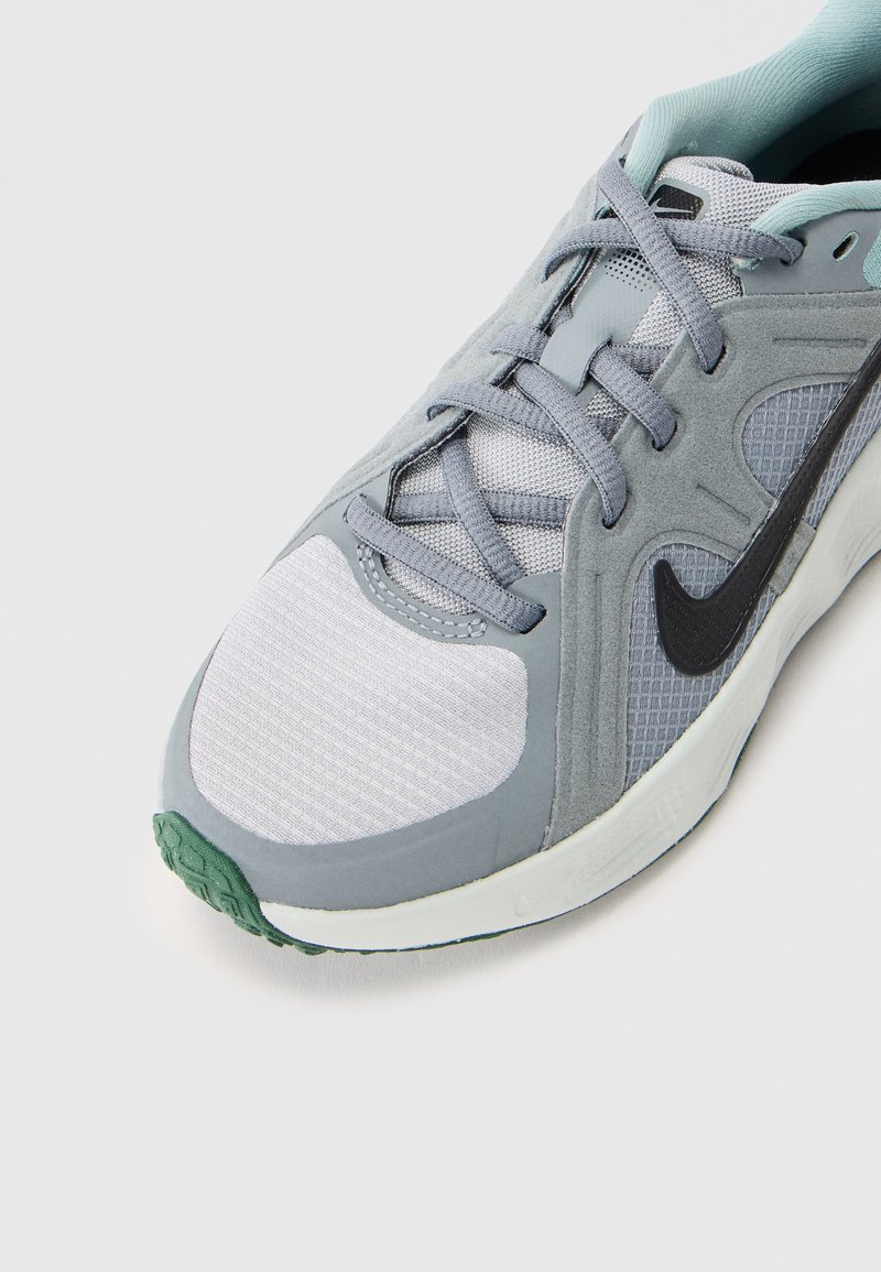 Zapatilla atlética gris con parte superior sintética que presenta texturas de malla y ante. Logotipo negro a un lado y suela de goma verde.