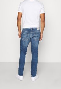 Vaqueros de mezclilla en azul medio con un corte slim, con pierna recta, bolsillos traseros tradicionales y un sutil detalle de desgaste. Combinados con una camiseta blanca.