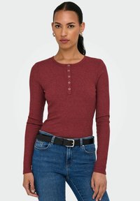 Ribbet, burgunder langermet topp med rund hals og fem knapper, kombinert med høy midje blå jeans og et svart belte.