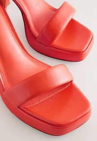 Sandali slingback in pelle rossa con cinturini larghi e piatti e design a punta quadrata. Il tacco è spesso, offrendo una base solida.