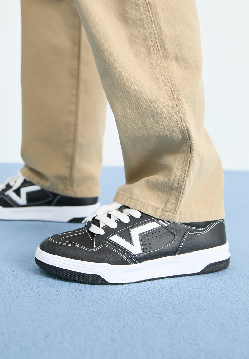 Des sneakers noir et blanc avec une tige en cuir texturé, un design rétro, des lacets blancs et une semelle en caoutchouc, associés à un pantalon beige clair.