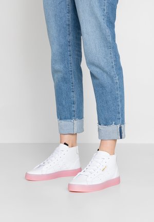 Personne portant un jean bleu clair retroussé et des baskets montantes blanches avec des semelles roses, debout sur un sol clair.