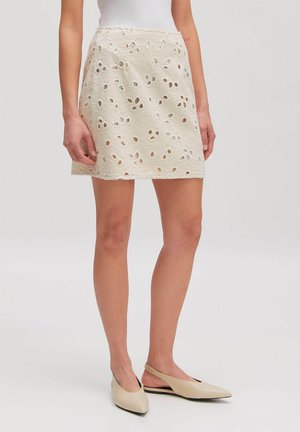 Frau trägt einen cremefarbenen Minirock aus Lochspitze und beige spitze Slingback-Ballerinas, stehend vor einem schlichten Hintergrund.