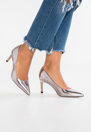 Zapatos de tacón alto de charol plateado con punta afilada y tacón stiletto de 8 cm. Llevados con jeans de mezclilla azul con dobladillo deshilachado.