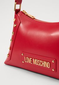 Rote Kunstleder-Handtasche mit goldfarbenen Beschlägen. Verfügt über Nieten an der Seite und einem Schild mit der Aufschrift "LOVE MOSCHINO". Mit Reißverschluss.