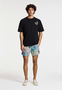 Fekete rövid ujjú póló mellkasi mintával, színes virágos fürdőnadrággal párosítva. A modell fehér cipőt visel.