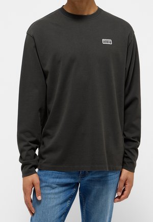 Person trägt ein schwarzes Langarmshirt von Mustang und blaue Jeans, mit einem kleinen "Mustang ESTD 1932" Logo auf der Brust.