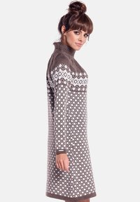 Kobieta z brązowymi włosami związanymi w kok, ubrana w długi sweterkowy brązowo-biały wzorzysty sweter z długim rękawem i wysokim kołnierzem, stojąca bokiem.