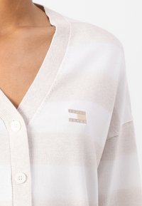Cárdigan de un beige claro con rayas blancas, con un escote en V, cuatro botones y un pequeño parche con el logo "Tommy Jeans" en el lado izquierdo del pecho.