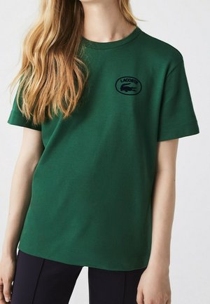 Mujer con camiseta de manga corta Lacoste verde oscuro con logo de cocodrilo negro en el pecho izquierdo, combinada con pantalones negros.
