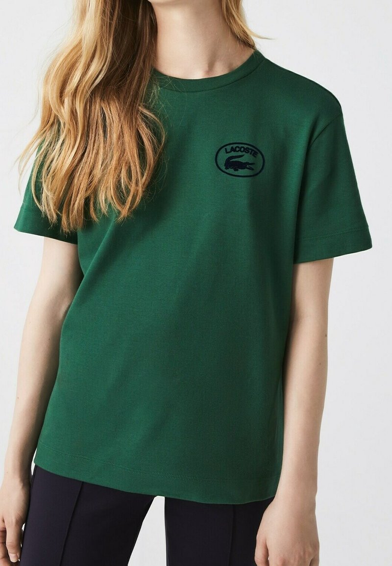 Femme portant un t-shirt Lacoste manches courtes vert foncé avec un logo crocodile noir sur le côté gauche de la poitrine, associé à un pantalon noir.