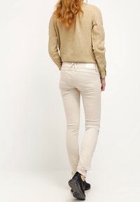 Jeans ajustados de color beige claro con una cintura de altura media, bolsillos traseros y una textura sutil. Combinados con una chaqueta corta de lino beige y zapatillas negras.
