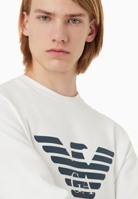 Felpa bianca con un emblema grafico in blu scuro che presenta strisce orizzontali e la scritta "GA" sotto. Texture liscia, design a collo rotondo.