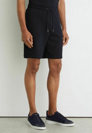 Mand iført sorte shorts med snøre og mørkeblå sneakers står op ad en hvid væg med hænderne afslappet ved siden.