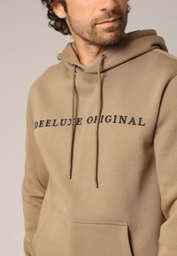 Bruine hoodie van zacht materiaal met een kangoeroezak, trekkoorden en "DEE LUXE ORIGINAL" in het zwart gedrukt op de borst.