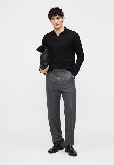 Άνδρας με μαύρο πουκάμισο με μακριά μανίκια και φερμουάρ στο γιακά και γκρι παντελόνι, κρατώντας μαύρη δερμάτινη τσάντα, στέκεται μπροστά σε απλό φωτεινό φόντο.