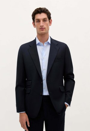 Marineblazer aus strukturiertem Stoff mit Reverskragen, zwei frontalen Taschen und einer einzelnen Knopfschließe. Leicht blaues Hemd darunter.