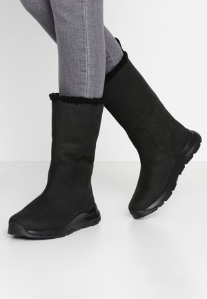 Persoon draagt zwarte winterlaarzen tot halverwege de kuit met bontvoering en grijze strakke jeans, staand op een witte vloer tegen een witte achtergrond.