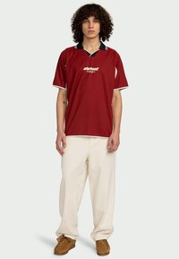 Mladá osoba s kučeravými vlasmi, oblečená v červenej polokošeli s krátkym rukávom, krémových širokých nohaviciach a hnedých mokasínach, stojaca čelom.