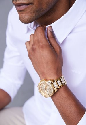 Man in white shirt adjusting collar, wearing a gold wristwatch with a diamond-studded band and detailed chronograph face.