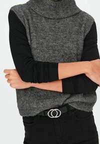 Personne portant un haut à manches longues noir sous un pull en tricot gris sans manches avec un col roulé, avec un pantalon noir et une ceinture à boucle double, les bras croisés.