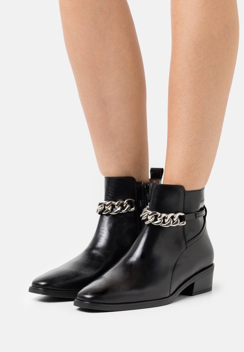 Jambes portant des bottines noires brillantes à talons bas avec un détail de chaîne argentée sur le devant, sur fond blanc uni.