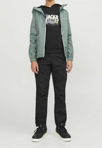 Giacca impermeabile verde chiaro con cappuccio, indossata sopra una felpa nera con cappuccio con scritte grafiche bianche, abbinata a pantaloni cargo neri e sneakers nere.