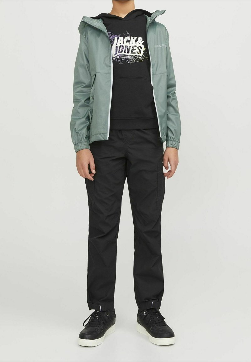 Giacca impermeabile verde chiaro con cappuccio, indossata sopra una felpa nera con cappuccio con scritte grafiche bianche, abbinata a pantaloni cargo neri e sneakers nere.