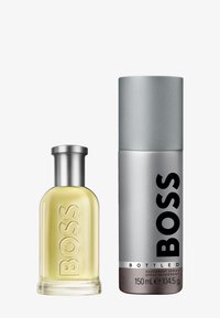 Szklana butelka żółtej wody toaletowej z srebrną nakrętką obok srebrnego pojemnika z dezodorantem z czarnym tekstem, oznaczonego "BOSS Bottled."