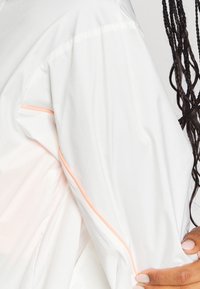 Valkoinen kevyt takki, jossa on sileä pinta ja persikan värinen reunus hihoissa ja olkasaumassa.