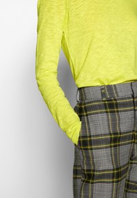 Chemise à manches longues de couleur jaune néon associée à un pantalon à carreaux gris avec des accents verts et jaunes, mettant en avant un design texturé.