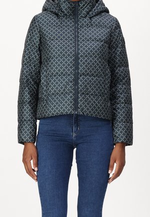 Woman wearing a navy blue quilted jacket with white diamond pattern and hood, paired with blue jeans, standing against a plain background.