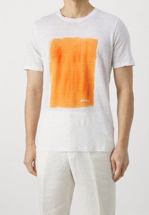 Homme portant un t-shirt blanc à manches courtes avec un grand graphisme rectangulaire orange sur le devant et un pantalon blanc sur un fond uni.