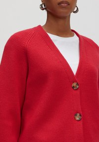 Rot gerippter Cardigan mit V-Ausschnitt und zwei großen Knöpfen. Das Design umfasst einen strukturierten Stoff und einen lockeren Schnitt, getragen über einem weißen Hemd.