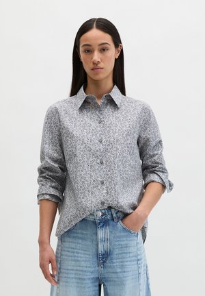 Frau mit glatten, langen dunklen Haaren trägt ein graues, geblümtes Hemd mit aufgekrempelten Ärmeln und hellblaue Jeans mit hoher Taille.