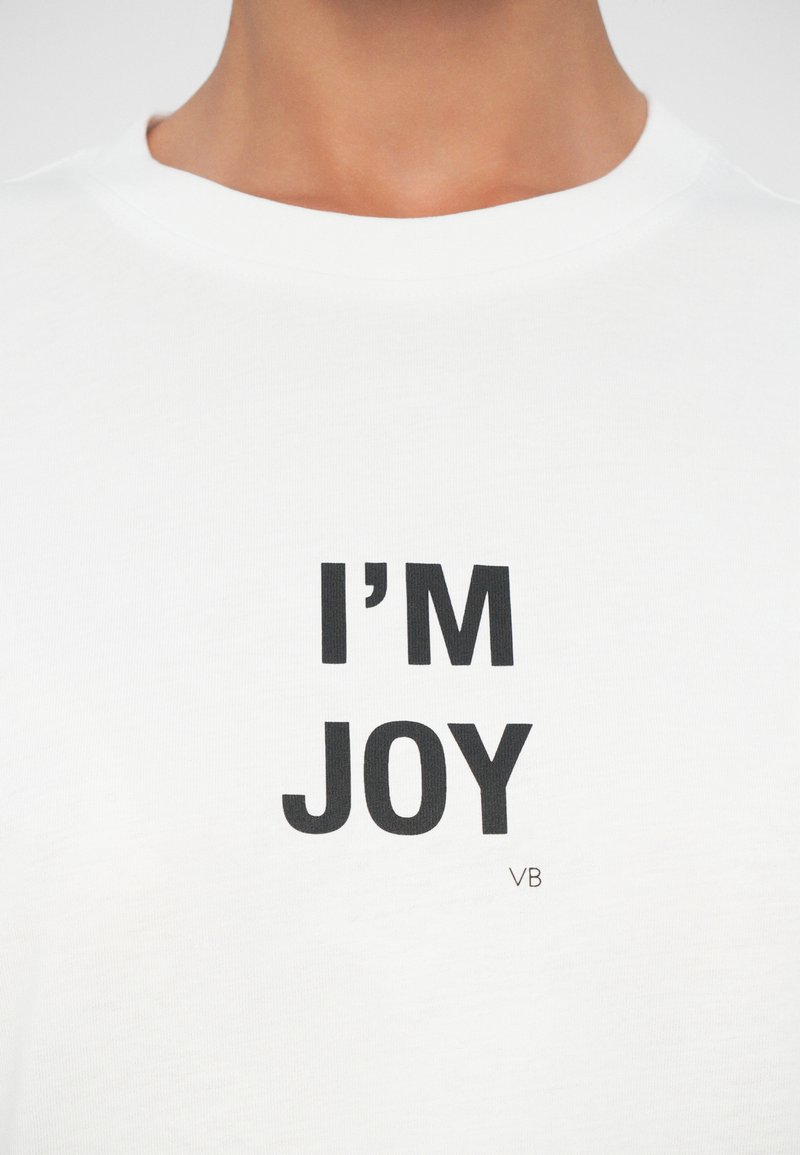 Vit bomulls t-shirt med fet svart text som säger "JAG ÄR GLÄDJE." Inkluderar en liten "VB"-logotyp nära botten. Minimalistisk design.