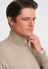 Beżowy sweter z dzianiny z kołnierzem typu golf; gładka faktura; model dotykający podbródka; noszący srebrny zegarek na nadgarstku.