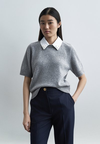 Jeune femme aux longs cheveux foncés portant un pull gris à manches courtes sur une chemise blanche à col, et un pantalon bleu marine, une main dans la poche.