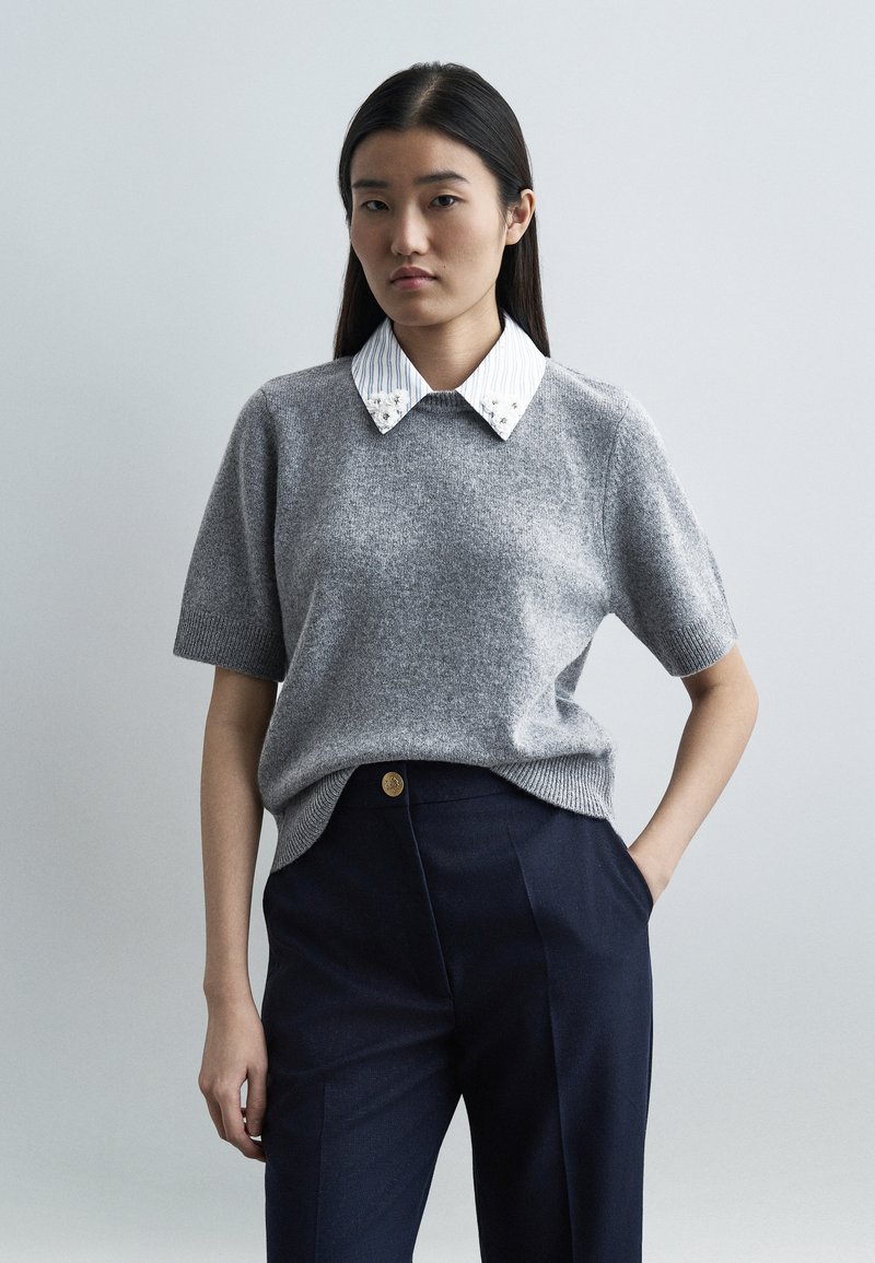 Jeune femme aux longs cheveux foncés portant un pull gris à manches courtes sur une chemise blanche à col, et un pantalon bleu marine, une main dans la poche.