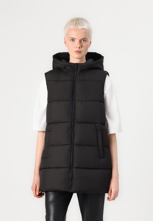 Sort polstret vest med hætte, der har fuld lynlås, sidelommer og quiltet tekstur. Båret over en hvid skjorte, parret med sorte bukser.