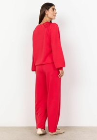 Roter Baumwoll-Sweatshirt mit langen Ärmeln und Spitzen-details auf der Rückseite, kombiniert mit lockeren roten Baumwollhosen. Flache beigefarbene Schuhe runden das Outfit ab.