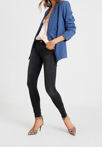 Mujer con una camisola rosa claro, blazer azul, jeans skinny negros y zapatos de tacón de serpiente, de pie con una mano en el bolsillo.