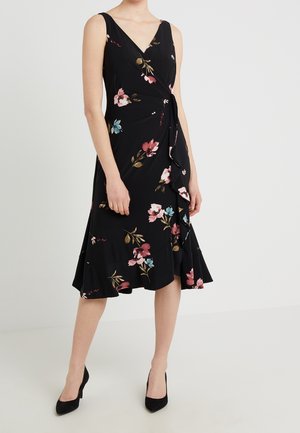 Frau trägt ein schwarzes ärmelloses Blumen-Wickelkleid mit Rüschensaum und schwarzen High Heels, steht vor einem schlichten hellen Hintergrund.