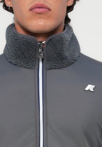 Grijze jas met een getextureerde fleece kraag, een ritssluiting aan de voorkant met oranje details en een logovoorziening op de borst.